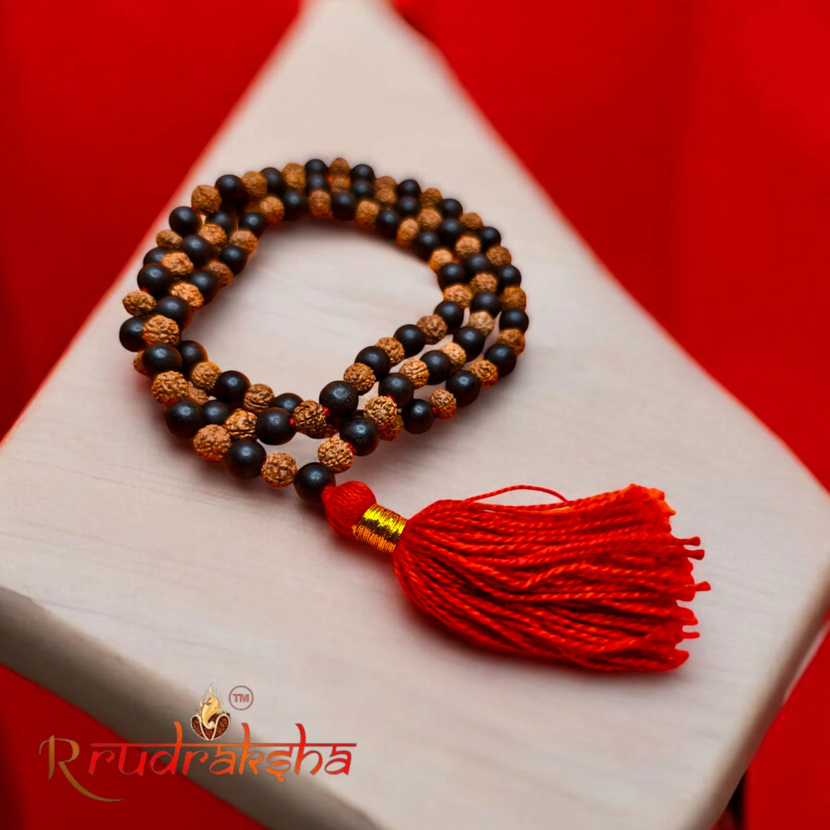 Mala necklace with Rudraksh and Karungali Malai beads and a red tassel on a white surface with a red background, featuring the R Rudraksha brand.