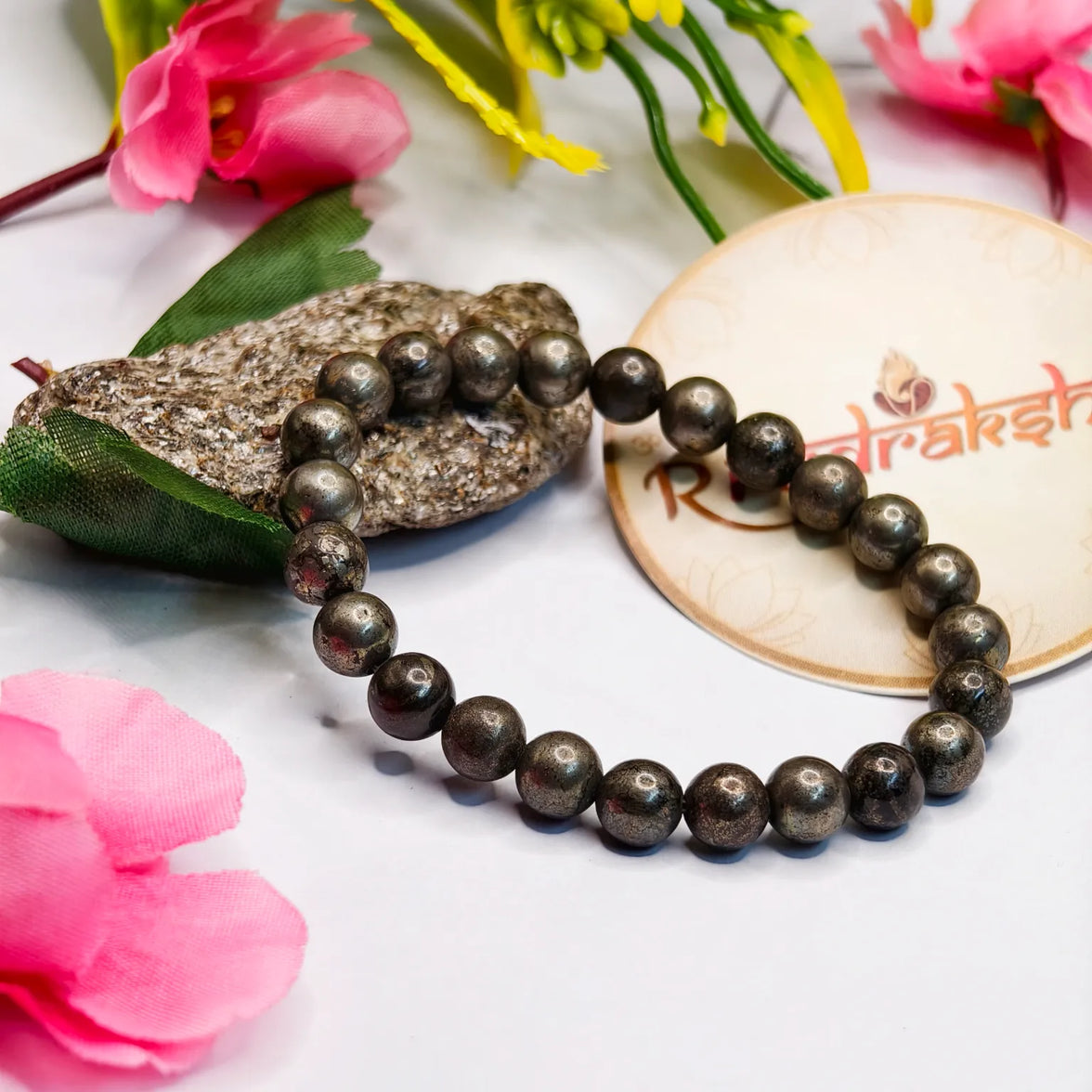 Pyrite bracelet Lab Certified product bracelet on a white surface with pink flowers and a stone. 