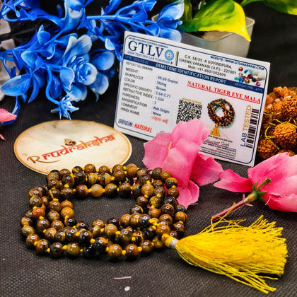 Tiger eye mala with lab identification report and Radhakrishna logo on a textured surface with flowers.